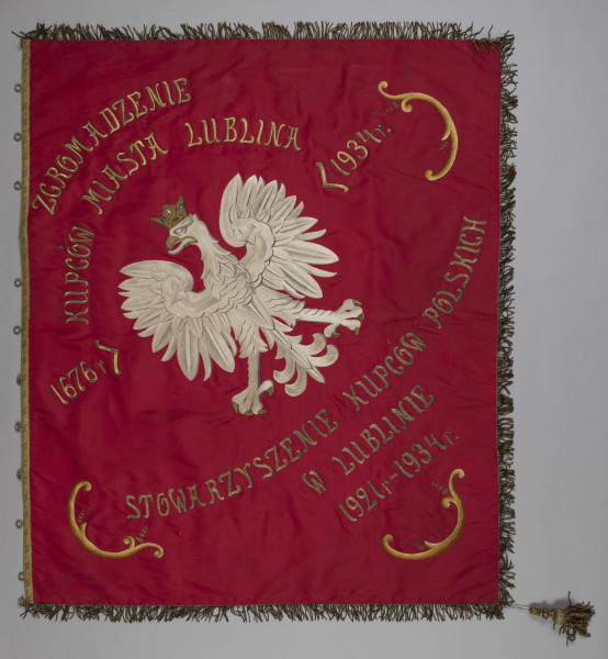 ML/H/301 - Sztandar Zgromadzenia Kupców miasta Lublina i Stowarzyszenia Kupców Polskich w Lublinie. Wzdłuż jednej z krawędzi ozdobne obustronne obszycie oraz metalowe okrągłe mocowania do przyczepienia na drzewcu. Wokół obszycie frędzlami. W jednym rogu umocowany chwost w kolorze frędzli. Na awersie pośrodku na czerwonym tle haftowany biały orzeł ze złotą koroną (ze sztucznymi kamieniami), wokół niego półkoliście ułożone haftowane złotawe napisy z nazwami dwóch organizacji oraz skrajnymi datami rozdzielonymi indeksami. Na awersie na kremowym tle wizerunek Matki Boskiej. Postać ukazana na stylizowanej granatowej sferze i złotym półksiężycu wspartymi na obłoku w geście błogosławieństwa. Twarz, dłonie i stopa malowane. Błękitne i czerwone szaty haftowane. Wokół sylwetki (głowy i rąk) rozchodzące się promienie (haftowane oraz wyszywane złotawymi cekinami). W narożach rozmieszczone haftowane barwne motywy kwiatowe (wyszywane fioletową, zieloną, czerwoną, fioletową oraz złotą nicią).  