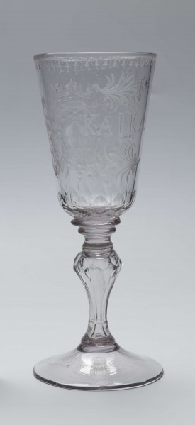 S/CS/244/ML - Stopa okrągła, lekko uniesiona, trzon tralkowy, z pęcherzykiem powietrza, fasetowany, od czaszy oddzielony pierścieniem i dwoma talerzykami, czasza stozkowata, u nasady zaokrąglona, szlifowana w dwa pionowe rzędy owalnych faset. Dekoracja rytowana: na czaszy dwustronna - z jednej strony owalna tarcza z parą gołąbków połączonych dziobkami stojących na serduszkach w rozbudowanym kartuszu z uszakami, z którego wyrastaja gałązki; po drugiej stronie litery 