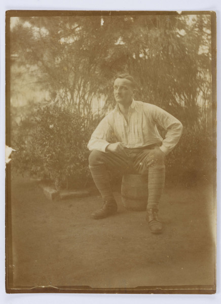 Fotografia przedstawia Jerzego Pola, siedzącego na beczce, z opartymi na kolanach rękoma. Mężczyzna w średnim wieku, ubrany w białą koszulę, ciemne spodnie wpuszczone w zawiązywane trzewiki. W tle widoczne liczne krzewy i drzewa. Fotografia obwiedziona białą ramką.