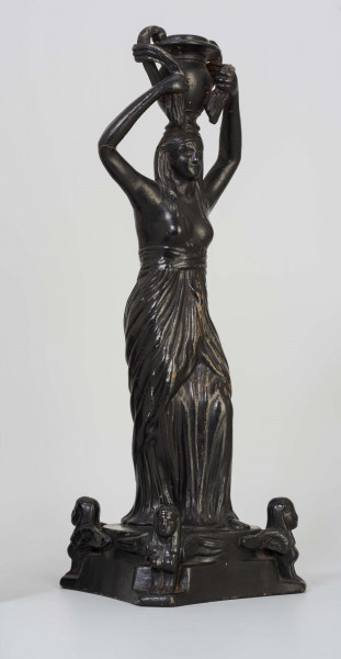 Świecznik figuralny z kariatydą; cokół czworoboczny górą przechodzący w koło, w narożach figurki sfinksów, na nim stojąca postać kobiety z wazonem na głowie, który podtrzymuje uniesionymi rękami. Kobieta do pasa naga, ubrana jest w fałdzistą szatę przwiązaną u góry, na głowie chusta opadajaca na ramiona. Całość malowana lakierem na czarno.
