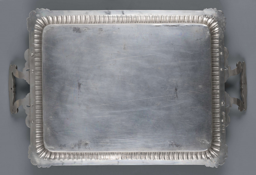 S/Mt/496/ML - Taca prostokątna o zaokrąglonych narożach, z dwoma uchwytami na krótszych bokach. Lustro gładkie, kołnierz podniesiony, trybowany w pionowe paski naprzemian szersze i węższe, brzeg obwiedziony sztancowaną taśmą z motywem palmetek, w narożach wzbogaconym maskami flankowanymi przez gryfy, uchwyty w formie  wolut połączonych toczoną tralką - w osadzeniach liście akantu. 
Na odwrocie sygnatura segmentowa:
- znak miejski Petersburga
- znak imienny złotnika  