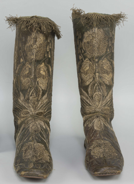 S/T/42/ML - Para butów z wysokimi cholewkami. Cholewka z miękkiej,zielonej skóry,  z dekoracyjnym haftem złotą nicią. Haft z dużym motywem kwiatowym: piwonie, kampanule i drobne kwiatki niezapominajki, tulipany(?), wydłużone liście i wici roślinne, wygięte łukowato.  Wewnatrz buty mają wykończenie białym, cienkim płótnem. Brzegi cholewki obszyte złota koronką z frędzlą. Podeszwy cieńkie z jednego kawałka skóry, bez obcasów, płaskie, z przodu prosto ścięte. Obydwie, skórzane podeszwy mają taki sam wykrój, bez rozróżnienia na lewą i prawą.
