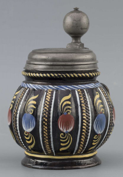 S/CS/595/ML - Kufel  kulisty, o niskiej konicznej stopie i niskiej szyi z pierścieniem, ucho taśmowe w kształcie „C”, pokrywka cynowa. Korpus pionowo kanelowany, podzielony na pasy na przemian gładkie i rytowane w ukośną kratkę. Pokryty szkliwem w kolorze ciemnego kobaltu, zdobiony malowanymi ornamentami podkreślającymi podziały powierzchni, w kolorach: czerwień, żółcień, błękit, biel. Pokrywka płaska, z pierścieniami i uchwytem kulkowym, na zawiasie umocowanym do ucha. Na wierzchu pokrywki grawerunek przedstawiający zawiły ornament – być może monogram spleciony z liter JGL – pod koroną, w uproszczonym wieńcu, z datą 1716.  