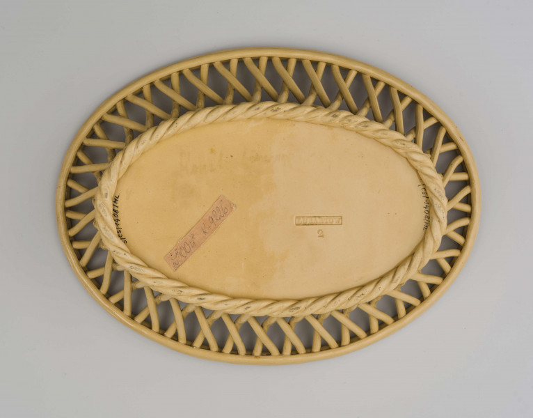 Koszyczek owalny głęboki; dno pełne od zewnątrz obwiedzione warkoczem, boki z okrągłych wałków przeplecionych ukośnie, połączonych profilowaną obręczą tworzącą krawędź. W lustrze (na dnie) dekoracja reliefowa: pośrodku bukiet kwiatów w wazonie, wokół brzegu wić laurowa z owocami. Całość pokryta połyskliwą glazurą w kolorze jasnego ugru.
 