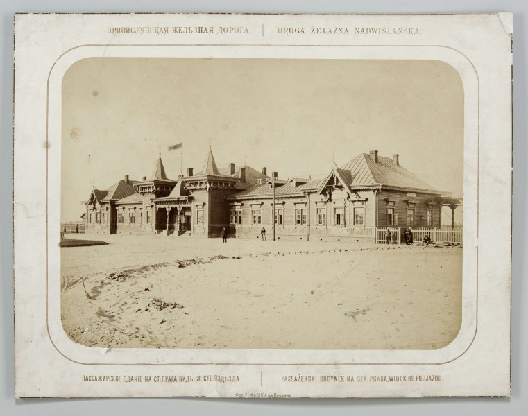 Budynek drewniany, parterowy, nakryty dachem z blachy. Wejście do budynku w formie podcienia wspartego na drewnianych filarach, do drzwi prowadzą schody. Niektóre elementy dachu w formie wieżyczek, nad wejściem umieszczona flaga. Szczyty i obramienia okien wykończone dekoracyjną formą stolarki. Teren częściowo uporządkowany, ogrodzony niskim płotem, przed budynkiem stoi kilka osób. Fotografia o zaokrąglonych rogach, obrysowana dookoła cienką ramką, naklejona na tekturę. Powyżej i pod fotografią napisy po rosyjsku - z lewej i po polsku - z prawej strony. W dolnej części informacja o autorze zdjęcia po rosyjsku.