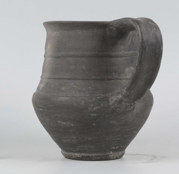 Dno płaskie, wyraźnie wyodrębnione stopką skośnie ściętą ku dołowi; dno wyodrębnione od części przydennej dookolnym wgłębieniem; lekko rozszerzające się ku górze, brzusiec łagodnie zaokraglony, ku górze zwężający się, przechodzący w wysoką, cylindryczną szyjkę; wylew płasko ścięty, rozchylony na zewnątrz; na największej wydętości brzuśca dookolny żłobek; w miejscu przejścia brzuśca w szyję i w połowie szyi dookolny wypukłe listwy; ucho z taśmy środkiem wklęsłej, biegnące od wylewu do załomu brzuśca, szyja zdobiona różnym ornamentem (trzy pasy) przez wygładzenie; powierzchnia gładzona, wyświecona
wymiary dodatkowe: 17,0 cm (obwód); 8,0 cm (śr. dna); 0,3 cm (wys. stopki); 0,5 cm (wys. szyi)