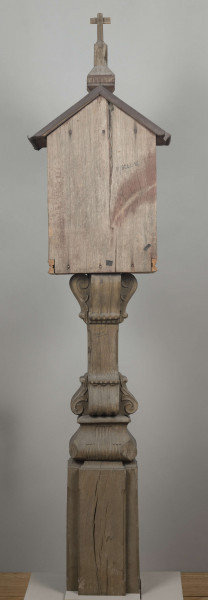 E/8062/1/ML - Kapliczka słupowa wykonana ręcznie siekierą i dłutami. Dębowy, profilowany ozdobnie słup w formie czterograniastej kolumny. Baza przy podstawie spróchniała, ma wgłębione narożniki. Żłobienia, głębokości 3,5 cm, uwypuklają płaszczyzny. Na przedniej ścianie wyryty napis: „Obrobił i postawił własnemi rencznie Jan Oliński dnia 17 marca 1933 r.”. Baza zakończona elementem o przekroju odwróconego trapezu wys. 17 cm i podstawie 26 cm, o zaokrąglonych dolnych narożnikach. Na nim trzon ozdobny czworograniasty, wys. 18 cm. Podstawa trzonu i głowica 4 esowatych żeberek, szerokości 12 cm. Na bocznych pełnych ściankach żeberek uproszony liść akantu. Na głowicach – kapliczka ażurowa. Kolumienki i szczyt brzozowe. Baza kolumny ma kształt graniastosłupa (wys. 12 cm, szer. 7 cm); trzon walcowaty (wys. 22 cm, szer. 8 cm), wybrzuszony, z pionowymi szerokimi rowkowaniami. Głowica (wys. 17 cm), utworzona z uproszczonych liści akantu i dwu złączonych w podstawy rozbieżnych wolut. Szczyt – tympanon ze śladami błękitnej polichromii, ozdobiony perliście małymi sześcianami o zaokrąglonych dolnych krawędziach. U szczytu drewniana kotara (lambrekin) z udrapowanych łukowatych żłobień, w środku ujęty prostą taśmą. Dach dwuspadowy, sosnowy o podciętych i podwiniętych krawędziach dolnych, pokryty blachą. Plecy i podstawa kapliczki – proste, sosnowe.