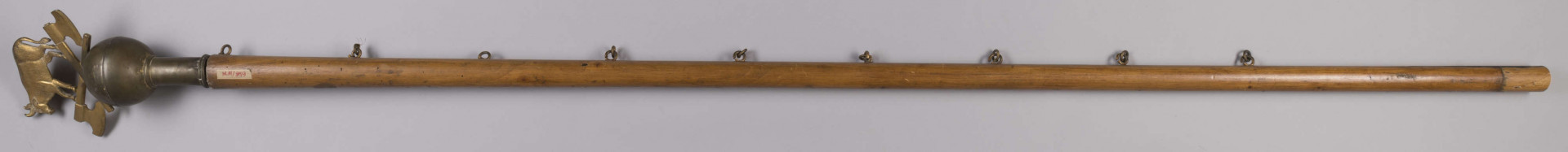 ML/H/959 - Sztandar Zgromadzenia Majstrów Rzeźniczych m. Lublina. Na awersie na amarantowym wzorzystym tle postać św. Jana Nepomucena w szatach pontyfikalnych, z krzyżem i gałązką palmową w dłoni oraz z Dzieciątkiem Jezus. W tle krajobraz z zabudowaniami i wieżami kościołów. Wokół haftowany ornament roślinny. Twarz i dłonie postaci malowane olejną farbą. Na rewersie haftowane złotą nicią napisy z nazwiskami starszego, podstarszego, podskarbiego cechu oraz ojca gospodniego. Nad nimi owieczka w pozycji leżącej ułożona na podstawie w formie krzyża. Wokół ornament kwiatowo-roślinny ze stylizowanymi liliami. Między ornamentami obu stron sztandaru wszyte ozdobne cekiny. Wokół sztandaru w formacie zbliżonym do kwadratu ozdobne frędzle. Po jednej stronie doszyte kółka do podtrzymywania sztandaru. Drzewiec zwieńczony blaszaną kulą ze skrzyżowanymi toporami oraz wołem pośrodku.