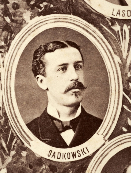 Fotografie w odcieniu sepii umieszczone na tle drzew i roślin, w centralnym miejscu postać kobiety ze sztandarem i aniołkami, wkomponowane widoki budowanej stacji, mostu kolejowego i lokomotywy. Największe zdjęcie w górnej, środkowej części bez podpisu, prawdopodobnie inż. głównej kolei nadwiślańskiej Tadeusz Chrzanowski lub jego następca Walery Titkow. Wśród podobizn pracownicy Biura Głównego Inżyniera: inż. Włodzimierz Stanisławski - pomocnik głównego inżyniera; inż. Feliks Kucharzewski -  pomocnik naczelnika biura; Feliks Kamocki - naczelnik kancelarii; Witold Lanci - naczelnik Wydz. Architektury; dr Wacław Lasocki- służba zdrowia; Władysław Radwan - sprawy zakupu gruntów; inż. Stanisław hr Scipio - projektant mostów kolejowych i jego współpracownicy: inż. Stefan Zieliński, inż. Maurycy Machalski, inż. Feliks Kucharzewski. Fotografia w passe - partout, za szkłem, oprawiona w drewnianą, profilowaną ramę.