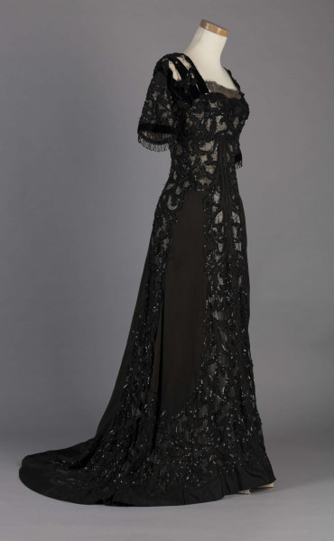 S/T/243/ML - Suknia wizytowa, czarna, uszyta z atłasu jedwabnego, jedwabnego tiulu i aksamitu, haftowana cekinami. Krótki rękaw z jedwabnego czarnego aksamitu, dekolt prostokatny, dość głęboki, wykończony wstawką haftowaną i aksamitem. Kolory są kontrastowe. Spodnia warstwa sukni, z białej tafty i muślinu, widoczna w partiach sukni szytych z czarnego tiulu, dodaje całości elegancji. Brzegi dekoracyjnej wici z tafty zaakcentowane naszytymi cekinami. Suknia do ziemi z tyłu krótki tren. Brak halki. 