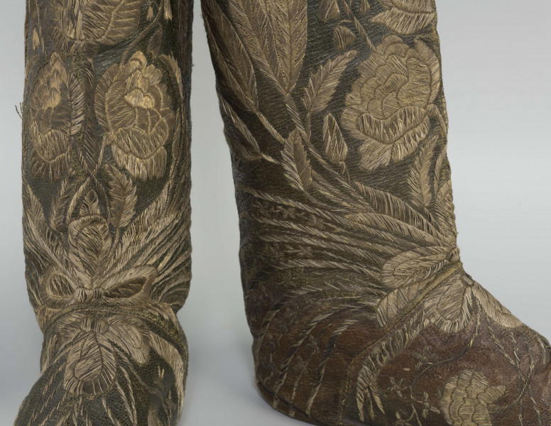 S/T/42/ML - Para butów z wysokimi cholewkami. Cholewka z miękkiej,zielonej skóry,  z dekoracyjnym haftem złotą nicią. Haft z dużym motywem kwiatowym: piwonie, kampanule i drobne kwiatki niezapominajki, tulipany(?), wydłużone liście i wici roślinne, wygięte łukowato.  Wewnatrz buty mają wykończenie białym, cienkim płótnem. Brzegi cholewki obszyte złota koronką z frędzlą. Podeszwy cieńkie z jednego kawałka skóry, bez obcasów, płaskie, z przodu prosto ścięte. Obydwie, skórzane podeszwy mają taki sam wykrój, bez rozróżnienia na lewą i prawą.