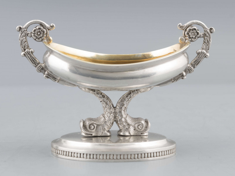 S/Mt/511/ML - Solniczka w formie łódki wspartej na dwóch delfinach, z owalną podstawą, z dwoma uchami z wici roślinnej. Brzeg czarki wywinięty, podstawa lekko podniesiona a jej brzeg zdobiony pionowym karbowaniem. Na spodzie sygnatura:
- imienna 