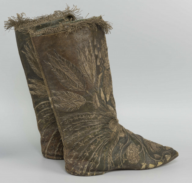 S/T/42/ML - Para butów z wysokimi cholewkami. Cholewka z miękkiej,zielonej skóry,  z dekoracyjnym haftem złotą nicią. Haft z dużym motywem kwiatowym: piwonie, kampanule i drobne kwiatki niezapominajki, tulipany(?), wydłużone liście i wici roślinne, wygięte łukowato.  Wewnatrz buty mają wykończenie białym, cienkim płótnem. Brzegi cholewki obszyte złota koronką z frędzlą. Podeszwy cieńkie z jednego kawałka skóry, bez obcasów, płaskie, z przodu prosto ścięte. Obydwie, skórzane podeszwy mają taki sam wykrój, bez rozróżnienia na lewą i prawą.
