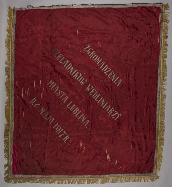 ML/H/960 - Sztandar Zgromadzenia Czeladników Wędliniarzy miasta Lublina. Dwustronnie opracowany, w kształcie prostokąta, obszyty frędzlami. Z jednej strony zaczepienia do mocowania na drzewcu. Na awersie, na czerwonym tle, w centralnej części postać św. Antoniego w habicie z kapturem, przepasanego sznurem i różańcem z czarnych koralików. Święty stoi na skale (?) wśród zieleni. W lewej dłoni trzyma w ręku figurę biczowanego Chrystusa przywiązanego do słupa. W prawej bicz (?) lub nieudolnie wyhaftowane litery „TAU” symbolizujące świętego. Wizerunek wykonany pełnym haftem, poza twarzą, dłońmi i stopami namalowanymi olejną farbą. Dookoła sceny w czterech rogach haftowane gałązki żółtych róż. Na rewersie na czerwonym tle obszytym pasem srebrnego galonu pośrodku haftowany napis z nazwą posiadacza oraz datą.