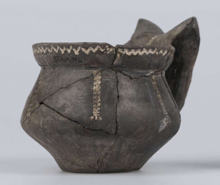 524/A/ML/2 - Kubek gliniany o niskiej szyjce, lejkowato rozchylonej o ukośnie ściętej do wewnątrz krawędzi, oddzielona ostrym załomem od dwustozkowatego brzusca, którego maksymalna wydętość przypada nieco poniżej połowy wysokości naczynia, dno silnie wyodrębnione, wklęsłe. Górny przyczep ucha ansa lunata przylega do krawędzi wylewu, dolny siega największej wydętości brzuśca. Ornament odciśniety prostokątnym stempelkiem, pod wylewem pozioma linia zygzakowata, na brzuścu 6 pionowych pasm ornamentu drabinkowego zakończonych 3 ukośnymi odciskami stempelka (sięgających od załomu szyjka/brzusiec po maksymalną wydętość brzuśca). Takie pasmo także na osi symetrii ucha, pozioma linia drabinkowa łączy skrzydełka ucha anasa lunata. Ornament inkrustowany białą masą. Wymiary: wysokość 7,4 cm; wysokość z uchem ok. 9,1 cm; średnica wylewu 8 cm; szyjka 7,1 cm; brzusiec 9,5 cm; dno 4,4 cm. Surowiec - glina z drobnym piaskiem. Barwa powierzchni zewnętrznej - szarobrunatna, powierzchni wewnętrznej brunatna. Przełom szary. Sposób wykonania - lepienie, powierzchnia gładzona do wybłyszczenia, zdobiona, dobry wypał. zabiegi konserwatorskie - oczyszczanie, sklejanie, wygipsowanie, ucho częściowo zrekonstruowane. Stan zachowania - średni.