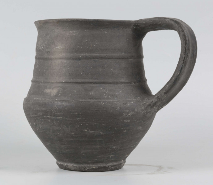 Dno płaskie, wyraźnie wyodrębnione stopką skośnie ściętą ku dołowi; dno wyodrębnione od części przydennej dookolnym wgłębieniem; lekko rozszerzające się ku górze, brzusiec łagodnie zaokraglony, ku górze zwężający się, przechodzący w wysoką, cylindryczną szyjkę; wylew płasko ścięty, rozchylony na zewnątrz; na największej wydętości brzuśca dookolny żłobek; w miejscu przejścia brzuśca w szyję i w połowie szyi dookolny wypukłe listwy; ucho z taśmy środkiem wklęsłej, biegnące od wylewu do załomu brzuśca, szyja zdobiona różnym ornamentem (trzy pasy) przez wygładzenie; powierzchnia gładzona, wyświecona
wymiary dodatkowe: 17,0 cm (obwód); 8,0 cm (śr. dna); 0,3 cm (wys. stopki); 0,5 cm (wys. szyi)