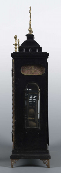 S/Mt/409/ML - Obudowa szafkowa, czworoboczna ze schodkowym zwieńczeniem, pokryta czarną politurą, oszklona z czterech stron, na czterech ażurowych nóżkach z mosiądzu; mosiężny uchwyt, ozdobny, składany a w górnych narożnikach sterczyny w formie płonących wazonów. Mechanizm chodu i bicia o napędzie sprężynowym, z wahadłem i dwoma dzwonkami; bicie godzin i kwadransów, budzik i repetier. Tarcza biało emaliowana z czarnymi cyframi arabskimi godzin i oznaczeniem minut, w niej trzy otwory na osie sprężyn; dwie ozdobne wskazówki. Powyżej tarczy wycięcie z widocznym wahadełkiem kontrolnym i trzy mniejsze emaliowane tarcze: środkowa z sygnaturą: Gottfried / Krohz / Cracovie, boczne z wyłączeniem chodu i bicia.