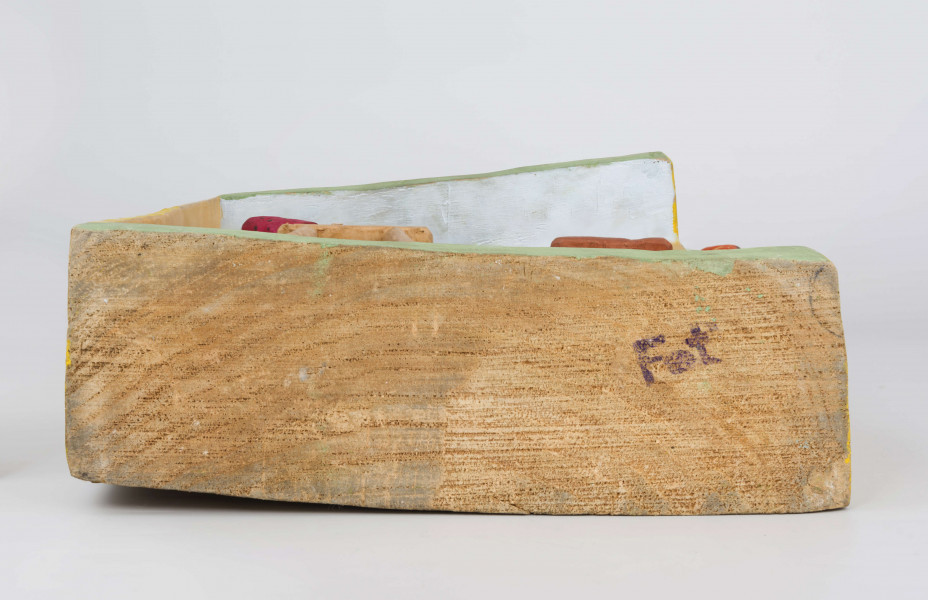 Płaskorzeźba w drewnie lipowym, polichromowana, przedstawiająca wnętrze kuchni, wykonana w jednym kawału drewna. Wnętrze kuchni zaznaczone z lewej strony występem szer. 6,7 cm i gł. 4,5 cm. U góry podkreślony „sufit” wys. 3 cm i gł. 5 cm. Na postumencie wys. 3,5 cm i gł. 4,5 cm. Z lewej strony zakomponowana postać kobiety (z profilu). Kobieta stoi lekko pochylona nad stołem, na którym wałkuje ciasto. Ubrana jest w długą czerwoną suknię w czarne kropki, z długim rękawem.  Na głowie ma zawiązaną, pod brodą, popielatą chustkę, spod której wystają czarne włosy. Twarz płaska, oczy lekko wypukłe, brwi czarne łukowate, nos długi prosty. 
Pod stołem stoi dzbanek, którego brzusiec ozdobiony jest czteropłatkowym kwiatem. Płatki w kolorze czerwony, środek zielony. Wylew podkreślony zielonym paskiem. Z prawej strony przy niskim brązowym stołku, po obu stronach stoją dzieci, które z jednej miski jedzą obiad. Z lewej strony stołka postać chłopca (wys. 12,5 cm) w szarych spodniach, sięgających za kolana i w zielonej koszuli wypuszczonej na spodnie. Twarz chłopca płaska, włosy czarne sięgające do ramion. W prawej ręce chłopiec trzyma łyżkę.  Z prawej – dziewczynka (wys. 13,6 cm), w długiej czerwonej sukience w żółto-czarne ciapki. Włosy czarne, sięgające ramion, twarz płaska. W prawej ręce trzyma łyżkę zanurzoną w misce, lewa opuszczona swobodnie. 
Na wys. 15,5 cm, z prawej strony, okno z kwiatami na parapecie i zasłonkami po bokach, do połowy okna. Wym. okna: wys. 14,5 cm, szer. 11,6 cm.