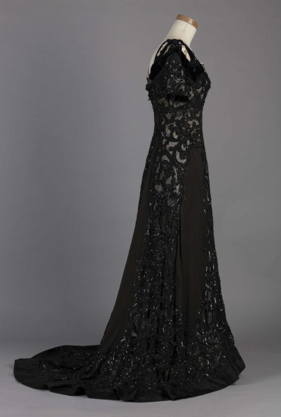 S/T/243/ML - Suknia wizytowa, czarna, uszyta z atłasu jedwabnego, jedwabnego tiulu i aksamitu, haftowana cekinami. Krótki rękaw z jedwabnego czarnego aksamitu, dekolt prostokatny, dość głęboki, wykończony wstawką haftowaną i aksamitem. Kolory są kontrastowe. Spodnia warstwa sukni, z białej tafty i muślinu, widoczna w partiach sukni szytych z czarnego tiulu, dodaje całości elegancji. Brzegi dekoracyjnej wici z tafty zaakcentowane naszytymi cekinami. Suknia do ziemi z tyłu krótki tren. Brak halki. 