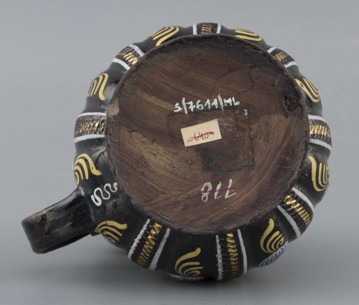 S/CS/595/ML - Kufel  kulisty, o niskiej konicznej stopie i niskiej szyi z pierścieniem, ucho taśmowe w kształcie „C”, pokrywka cynowa. Korpus pionowo kanelowany, podzielony na pasy na przemian gładkie i rytowane w ukośną kratkę. Pokryty szkliwem w kolorze ciemnego kobaltu, zdobiony malowanymi ornamentami podkreślającymi podziały powierzchni, w kolorach: czerwień, żółcień, błękit, biel. Pokrywka płaska, z pierścieniami i uchwytem kulkowym, na zawiasie umocowanym do ucha. Na wierzchu pokrywki grawerunek przedstawiający zawiły ornament – być może monogram spleciony z liter JGL – pod koroną, w uproszczonym wieńcu, z datą 1716.  