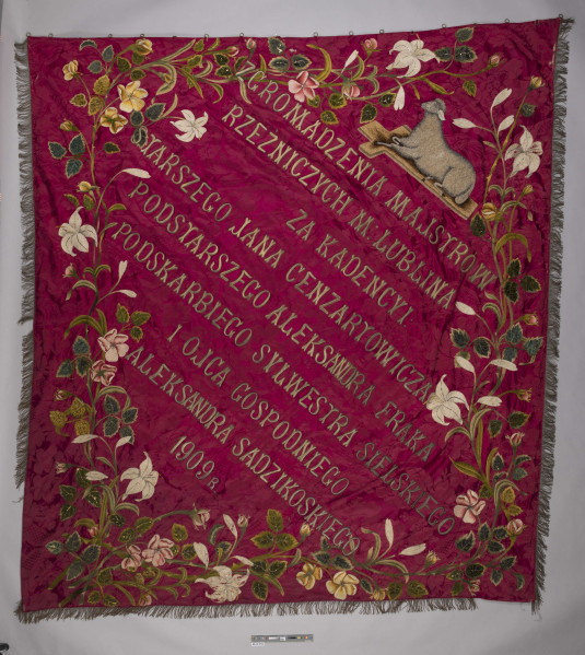 ML/H/959 - Sztandar Zgromadzenia Majstrów Rzeźniczych m. Lublina. Na awersie na amarantowym wzorzystym tle postać św. Jana Nepomucena w szatach pontyfikalnych, z krzyżem i gałązką palmową w dłoni oraz z Dzieciątkiem Jezus. W tle krajobraz z zabudowaniami i wieżami kościołów. Wokół haftowany ornament roślinny. Twarz i dłonie postaci malowane olejną farbą. Na rewersie haftowane złotą nicią napisy z nazwiskami starszego, podstarszego, podskarbiego cechu oraz ojca gospodniego. Nad nimi owieczka w pozycji leżącej ułożona na podstawie w formie krzyża. Wokół ornament kwiatowo-roślinny ze stylizowanymi liliami. Między ornamentami obu stron sztandaru wszyte ozdobne cekiny. Wokół sztandaru w formacie zbliżonym do kwadratu ozdobne frędzle. Po jednej stronie doszyte kółka do podtrzymywania sztandaru. Drzewiec zwieńczony blaszaną kulą ze skrzyżowanymi toporami oraz wołem pośrodku.