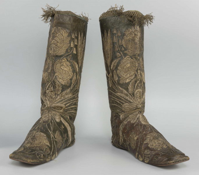 S/T/42/ML - Para butów z wysokimi cholewkami. Cholewka z miękkiej,zielonej skóry,  z dekoracyjnym haftem złotą nicią. Haft z dużym motywem kwiatowym: piwonie, kampanule i drobne kwiatki niezapominajki, tulipany(?), wydłużone liście i wici roślinne, wygięte łukowato.  Wewnatrz buty mają wykończenie białym, cienkim płótnem. Brzegi cholewki obszyte złota koronką z frędzlą. Podeszwy cieńkie z jednego kawałka skóry, bez obcasów, płaskie, z przodu prosto ścięte. Obydwie, skórzane podeszwy mają taki sam wykrój, bez rozróżnienia na lewą i prawą.