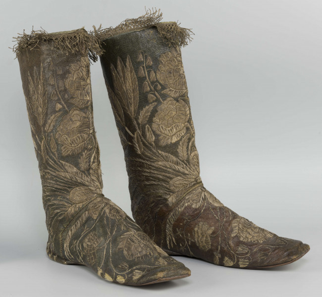S/T/42/ML - Para butów z wysokimi cholewkami. Cholewka z miękkiej,zielonej skóry,  z dekoracyjnym haftem złotą nicią. Haft z dużym motywem kwiatowym: piwonie, kampanule i drobne kwiatki niezapominajki, tulipany(?), wydłużone liście i wici roślinne, wygięte łukowato.  Wewnatrz buty mają wykończenie białym, cienkim płótnem. Brzegi cholewki obszyte złota koronką z frędzlą. Podeszwy cieńkie z jednego kawałka skóry, bez obcasów, płaskie, z przodu prosto ścięte. Obydwie, skórzane podeszwy mają taki sam wykrój, bez rozróżnienia na lewą i prawą.
