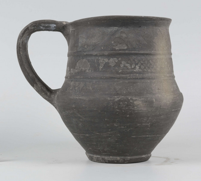 Dno płaskie, wyraźnie wyodrębnione stopką skośnie ściętą ku dołowi; dno wyodrębnione od części przydennej dookolnym wgłębieniem; lekko rozszerzające się ku górze, brzusiec łagodnie zaokraglony, ku górze zwężający się, przechodzący w wysoką, cylindryczną szyjkę; wylew płasko ścięty, rozchylony na zewnątrz; na największej wydętości brzuśca dookolny żłobek; w miejscu przejścia brzuśca w szyję i w połowie szyi dookolny wypukłe listwy; ucho z taśmy środkiem wklęsłej, biegnące od wylewu do załomu brzuśca, szyja zdobiona różnym ornamentem (trzy pasy) przez wygładzenie; powierzchnia gładzona, wyświecona
wymiary dodatkowe: 17,0 cm (obwód); 8,0 cm (śr. dna); 0,3 cm (wys. stopki); 0,5 cm (wys. szyi)