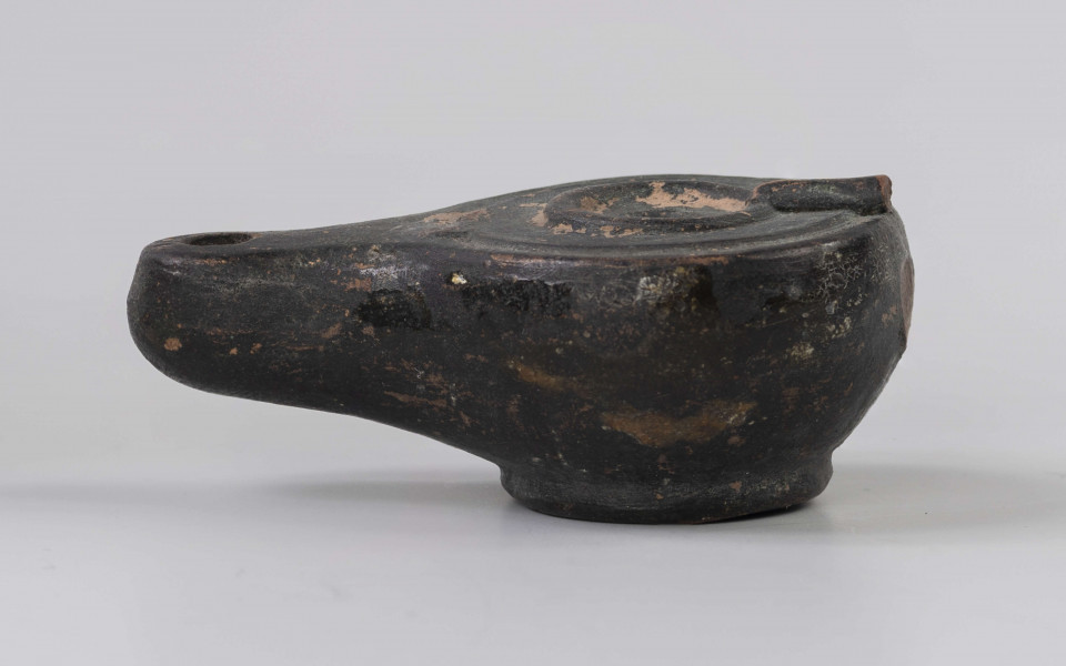 720/A/ML/2 - Lampka oliwna z utłuczonym uszkiem; mały otwór do wlewania oliwy obwiedziony plastycznym wałeczkiem, który jest połączony z uszkiem plastycznym wałeczkiem rozszerzającym się w kierunku uszka; wokół krawędzi dookolny złobek; mały otwór na płomien na wysmukłym, dość mocno wyciągniętym dziubku; dno wyodrębnione, wklęsłe; prawdopodobnie miniatura kultowej, pannońska wzorowana na lampach brązowych