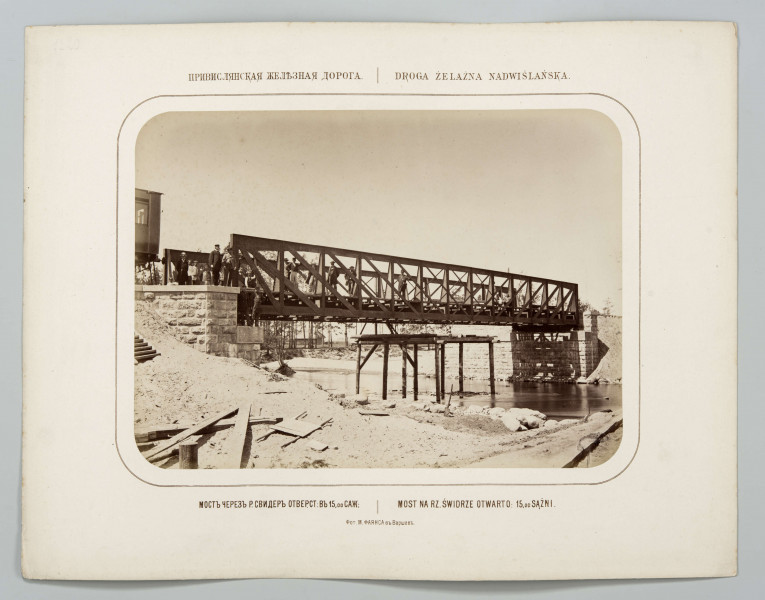 Fotografia przedstawia otwarcie mostu na rzece Świdrze. Na piaszczystym brzegu porozrzucane deski. Po lewej stronie na nasypie fragment wagonu i stojący mężczyźni. Most żelazny, kratowy, jednoprzęsłowy, 15 sążni. Pod mostem stoi wbita w dno rzeki drewniana konstrukcja. W tle drzewa i słabo widoczna stodoła. Fotografia o zaokrąglonych rogach, obrysowana dookoła cienką ramką, naklejona na tekturę. Powyżej i pod fotografią napisy po rosyjsku - z lewej i po polsku - z prawej strony. W dolnej części informacja o autorze zdjęcia po rosyjsku.