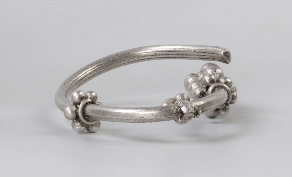 169/A/ML/1 - Zausznica srebrna składa się z kabłąka, na którym zawieszono wisiorek o winogronowatym kształcie. Ozdoby tego typu charakteryzują się kabłąkiem o jednym końcu ucięty prosto, drugim zaś zwiniętym w oczko. W połowie wysokości kabłąka znajdują się wieńce wykonane z granulek ograniczonych pierścieniami z drutu. W dolnej części kabłąka występuje wisiorek uformowany z kilku wieńców granulek zwieńczony granulką osadzoną na pierścieniu z drutu. 