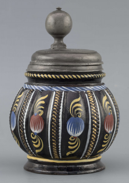 S/CS/595/ML - Kufel  kulisty, o niskiej konicznej stopie i niskiej szyi z pierścieniem, ucho taśmowe w kształcie „C”, pokrywka cynowa. Korpus pionowo kanelowany, podzielony na pasy na przemian gładkie i rytowane w ukośną kratkę. Pokryty szkliwem w kolorze ciemnego kobaltu, zdobiony malowanymi ornamentami podkreślającymi podziały powierzchni, w kolorach: czerwień, żółcień, błękit, biel. Pokrywka płaska, z pierścieniami i uchwytem kulkowym, na zawiasie umocowanym do ucha. Na wierzchu pokrywki grawerunek przedstawiający zawiły ornament – być może monogram spleciony z liter JGL – pod koroną, w uproszczonym wieńcu, z datą 1716.  