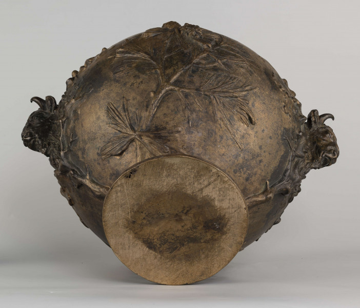 S/Mt/427/ML - Waza w kształcie spłaszczonej kuli, z niską, kolistą stopą i krótką szyją o lekko wywiniętym brzegu. Powierzchnia zdobiona reliefem: po bokach dwa uchwyty w formie głów żubrów, wokół nich gałązki dębu, pośrodku korpusu gałązki sosny z szyszkami wyrastające ze stopy. U góry na brzuścu rytowany napis majuskułą 