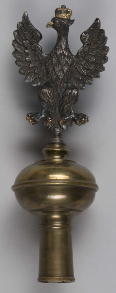 ML/H/200 - Sztandar Przedszkola i Seminarium Ochroniarskiego Marii Papiewskiej. Sztandar w formie kwadratu zszytego z dwóch kawałków ozdobnej tkaniny, obszyty frędzlami. Wzdłuż jednej z krawędzi mocowania w formie troczków do zaczepienia na drzewcu. Na awersie pośrodku tkaniny w kremowym kolorze w kwiatowe motywy herb miasta Lublina (koziołek wsparty o winną latorośl umieszczony na czerwonym polu na tarczy zwieńczonej koroną) oraz haftowany w półkolu czerwoną nicią napis z nazwą posiadacza. Na rewersie pośrodku wzorzystej tkaniny w kolorze czerwonym haftowany biały orzeł w złotej koronie wysadzanej klejnotami z umieszczoną poniżej haftowaną w kolorze złotym datą 1916 r. Metalowa głowica w kształcie orła w koronie umocowanego na kuli. Dodatkowo materiałowy pokrowiec zapinany u góry na napy i haftki, w kolorze zielonkawym, z wyhaftowanym pośrodku napisem w kolorze żółtym.