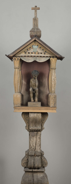 E/8062/1/ML - Kapliczka słupowa wykonana ręcznie siekierą i dłutami. Dębowy, profilowany ozdobnie słup w formie czterograniastej kolumny. Baza przy podstawie spróchniała, ma wgłębione narożniki. Żłobienia, głębokości 3,5 cm, uwypuklają płaszczyzny. Na przedniej ścianie wyryty napis: „Obrobił i postawił własnemi rencznie Jan Oliński dnia 17 marca 1933 r.”. Baza zakończona elementem o przekroju odwróconego trapezu wys. 17 cm i podstawie 26 cm, o zaokrąglonych dolnych narożnikach. Na nim trzon ozdobny czworograniasty, wys. 18 cm. Podstawa trzonu i głowica 4 esowatych żeberek, szerokości 12 cm. Na bocznych pełnych ściankach żeberek uproszony liść akantu. Na głowicach – kapliczka ażurowa. Kolumienki i szczyt brzozowe. Baza kolumny ma kształt graniastosłupa (wys. 12 cm, szer. 7 cm); trzon walcowaty (wys. 22 cm, szer. 8 cm), wybrzuszony, z pionowymi szerokimi rowkowaniami. Głowica (wys. 17 cm), utworzona z uproszczonych liści akantu i dwu złączonych w podstawy rozbieżnych wolut. Szczyt – tympanon ze śladami błękitnej polichromii, ozdobiony perliście małymi sześcianami o zaokrąglonych dolnych krawędziach. U szczytu drewniana kotara (lambrekin) z udrapowanych łukowatych żłobień, w środku ujęty prostą taśmą. Dach dwuspadowy, sosnowy o podciętych i podwiniętych krawędziach dolnych, pokryty blachą. Plecy i podstawa kapliczki – proste, sosnowe.