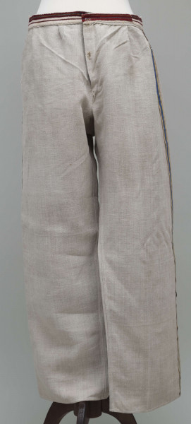 E/12943/ML - Spodnie z lnianego materiału tkanego splotem prostym. Szyte ręcznie i na maszynie. Nogawki szerokie ujęte w pasek szer. 3 cm, wzmocniony i ozdobiony paskiem płótna z ornamentem tkackim „pereborami”. Poniżej paska wszyte 2 trójkątne kliny oraz podobny w rozporku, zapinanym na 2 guziki. Na szwach bocznych nogawek naszyte kolorowe, fabryczne tasiemki, szer. 1,5 cm – żółta i niebieska.