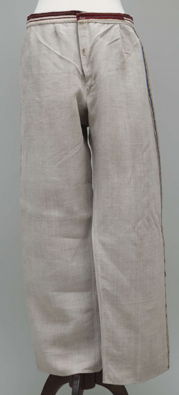 E/12943/ML - Spodnie z lnianego materiału tkanego splotem prostym. Szyte ręcznie i na maszynie. Nogawki szerokie ujęte w pasek szer. 3 cm, wzmocniony i ozdobiony paskiem płótna z ornamentem tkackim „pereborami”. Poniżej paska wszyte 2 trójkątne kliny oraz podobny w rozporku, zapinanym na 2 guziki. Na szwach bocznych nogawek naszyte kolorowe, fabryczne tasiemki, szer. 1,5 cm – żółta i niebieska.