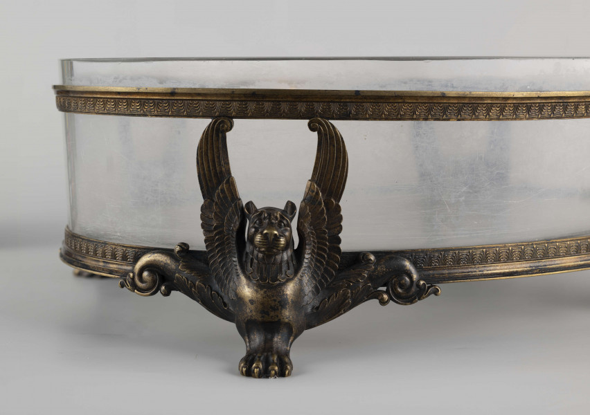 S/CS/1063/ML - Naczynie ze szkła bezbarwnego, grubościennego, owalne, z płaskim dnem, w oprawie metalowej złożonej z dwóch obręczy połączonych plastycznymi figurkami gryfów na nóżkach. Powierzchnia obręczy zdobiona plastycznym fryzem antykizujacych palmet. Oprawa metalowa patynowana. 
 