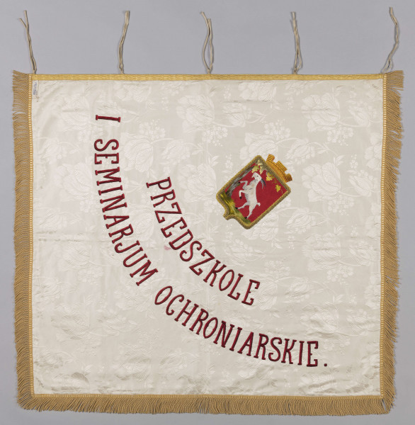 ML/H/200 - Sztandar Przedszkola i Seminarium Ochroniarskiego Marii Papiewskiej. Sztandar w formie kwadratu zszytego z dwóch kawałków ozdobnej tkaniny, obszyty frędzlami. Wzdłuż jednej z krawędzi mocowania w formie troczków do zaczepienia na drzewcu. Na awersie pośrodku tkaniny w kremowym kolorze w kwiatowe motywy herb miasta Lublina (koziołek wsparty o winną latorośl umieszczony na czerwonym polu na tarczy zwieńczonej koroną) oraz haftowany w półkolu czerwoną nicią napis z nazwą posiadacza. Na rewersie pośrodku wzorzystej tkaniny w kolorze czerwonym haftowany biały orzeł w złotej koronie wysadzanej klejnotami z umieszczoną poniżej haftowaną w kolorze złotym datą 1916 r. Metalowa głowica w kształcie orła w koronie umocowanego na kuli. Dodatkowo materiałowy pokrowiec zapinany u góry na napy i haftki, w kolorze zielonkawym, z wyhaftowanym pośrodku napisem w kolorze żółtym.