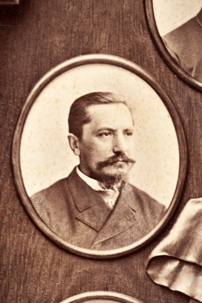 Pośrodku prostokątnego pola o zaokrąglonych narożnikach większa fotografia przyozdobiona wstęgą i gałązkami wawrzynu. Dookoła szesnaście symetrycznie rozmieszczonych mniejszych fotografii. W prawym górnym rogu dr Wacław Lasocki, naczelny lekarz kolei nadwiślańskiej w latach 1875 - 78. W polu ramy w górnej części medalion z płaskorzeźbionym popiersiem z profilu. Przedstawia Leopolda Kronenberga - ojca. Po bokach dwia półleżące aniołki trzymają wieniec laurowy. Po lewej stronie ramy postać robotnika Poleszuka, po prawej pracownika warsztatów kolejowych. W dolnej części front lokomotywy i złoty żeton uprawniający do bezpłatnej jazdy pociągiem I klasy. Fotografia naklejona na tekturę, w dolnej części u dołu sucha pieczęć zakładu fotograficznego.