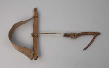 Szelki do niewodu (3 szt.) - Ujęcie z przodu pierwszych szelek z góry. Sprzęt pomocniczy ułatwiający ręczne wybieranie niewodu. Składają się z: parcianego pasa, będącego częścią pracującą drewnianego orczyka oraz krótkiego sznura,  do którego przywiązany jest fragment rozwidlonej gałęzi. Pas wykonany jest z jednego kawałka materiału, zszytego maszynowo. Na jego końcach wszyte są drewienka o owalnym przekroju, przy których znajdują się wycięte w materiale otwory. W jednym z nich umieszczona jest niewielka, wykonana ze sznurka pętla, w drugim sznurek łączący pas z orczykiem. Orczyk lekko wygięty, przy jednym końcu zwężony, przy drugim mający wcięcie o nieregularnym kształcie. W środkowej jego części znajduje się otwór przez który przechodzi sznurek, do którego przywiązana jest gałąź. Szelki znakowane są wyciętym w drewnie, niezidentyfikowanym merkiem oraz inicjałami: WM.