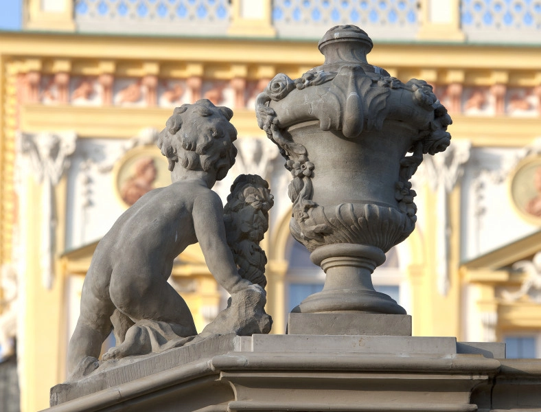 ujęcie od strony trzeciej. Putto z lewej, wazon z prawej. Putto ustawione na pochyłej balustradzie schodów, wazon na poziomej. Chłopiec ma tułów silnie skręcony w prawą stronę i jest zwrócone plecami do widza. Putto oparte na prawym kolanie, lewa noga wyprostowana. Prawa ręka wyprostowana wsparta na podstawie, w lewej ręce girlanda, tułów silnie skręcony w prawą stronę. Obok waza o kielichowatym kształcie dekorowana girlandami kwiatów i stylizowanymi liśćmi. Obie figury w kolorze szarobeżowym. W tle pałac.