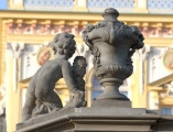 ujęcie od strony trzeciej. Putto z lewej, wazon z prawej. Putto ustawione na pochyłej balustradzie schodów, wazon na poziomej. Chłopiec ma tułów silnie skręcony w prawą stronę i jest zwrócone plecami do widza. Putto oparte na prawym kolanie, lewa noga wyprostowana. Prawa ręka wyprostowana wsparta na podstawie, w lewej ręce girlanda, tułów silnie skręcony w prawą stronę. Obok waza o kielichowatym kształcie dekorowana girlandami kwiatów i stylizowanymi liśćmi. Obie figury w kolorze szarobeżowym. W tle pałac.