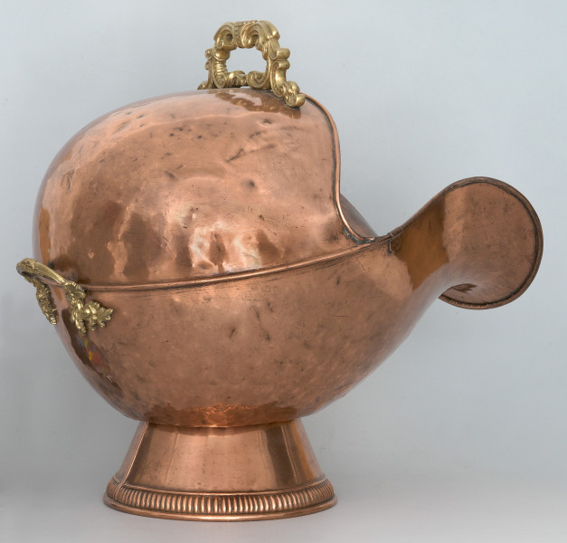 Ujęcie 3/4. Widok na boku i lekko z tyłu. Obiekt umieszczony na stopce o kształcie ściętego stożka. Pojemnik ma kształty zaokrąglone, z szerokim otworem o mocno wygiętej dolnej krawędzi po prawej stronie. Pojemnik i podstawa wykonane z miedzi, o kolorze jasnobrązowym. Na górze, przy krawędzi, oraz z tyłu widoczne rączki w kolorze złotym.