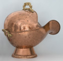 Ujęcie 3/4. Widok na boku i lekko z tyłu. Obiekt umieszczony na stopce o kształcie ściętego stożka. Pojemnik ma kształty zaokrąglone, z szerokim otworem o mocno wygiętej dolnej krawędzi po prawej stronie. Pojemnik i podstawa wykonane z miedzi, o kolorze jasnobrązowym. Na górze, przy krawędzi, oraz z tyłu widoczne rączki w kolorze złotym.