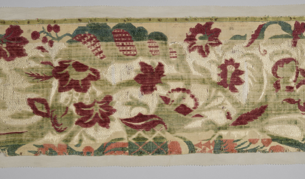 Pas tkaniny ułożony poziomo – zbliżenie na fragment. Widoczne motywy roślinne: czerwone kwiaty poprzeplatane zielonymi liśćmi na beżowym tle. W centralnej części szeroki pas tkaniny jest odbarwiony, liście są jaśniejsze, w tle jaśniejsze przeszycia. Wąski pas na dole również odbarwiony, motywy kwiatowe wpadają w pomarańcz. bryt Wil.Wn.125/1 - fragment, awers; ściana południowo-wschodnia