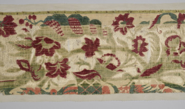 Pas tkaniny ułożony poziomo – zbliżenie na fragment. Widoczne motywy roślinne: czerwone kwiaty poprzeplatane zielonymi liśćmi na beżowym tle. W centralnej części szeroki pas tkaniny jest odbarwiony, liście są jaśniejsze, w tle jaśniejsze przeszycia. Wąski pas na dole również odbarwiony, motywy kwiatowe wpadają w pomarańcz. bryt Wil.Wn.125/1 - fragment, awers; ściana południowo-wschodnia