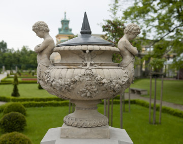 ujęcie od strony pierwszej. Wazon dekoracyjny na wysokim trzonie, szeroki z wywiniętą krawędzią. Ustawiony na wysokiej podstawie. Kolor beżowy. Dookoła brzuśca kwiatowa girlanda. Dwa symetryczne uchwyty w kształcie młodzieńców z podwójnymi rybimi ogonami – po lewej i prawej stronie. Przy szyjce ptak z rozpostartymi skrzydłami. Wazon przykryty metalowym hełmem ze szpiczastym uchwytem. W tle widoczny park i fragment pałacu.