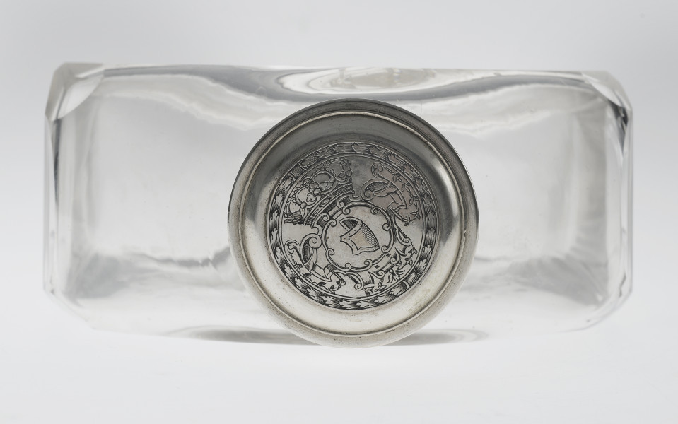 Ujęcie z góry. Na pierwszym planie okrągła, srebrna nakrętka z wygrawerowanym herbem Janina – wygiętą tarczą skierowaną w prawo. Nad herbem korona królewska, a całość otoczona wieńcem. Sama nakrętka przekręcona jest lekko w lewo. Na drugim planie widoczna szeroka, płaska butla o płaskich i przezroczystych ściankach.