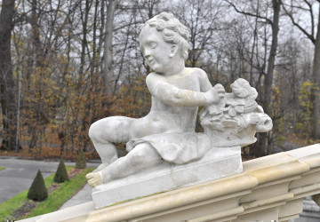 ujęcie od strony pierwszej. Nagie i pulchne dziecko siedzi z w pozycji półleżącej z nogami skierowanymi w lewą stronę. Z prawej  strony przytrzymuje ono kosz z kwiatami. Włosy dziecka są krótki i kręcone.